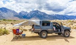 A frustrated overlander looking at a flat tire and a messy truck bed camping setup on a dirt road.