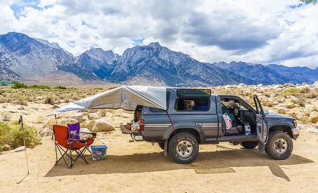 A frustrated overlander looking at a flat tire and a messy truck bed camping setup on a dirt road.