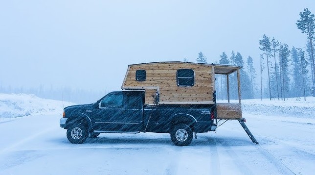 A truck parked in the snow with cozy, warm lighting glowing from the truck bed camper shell.